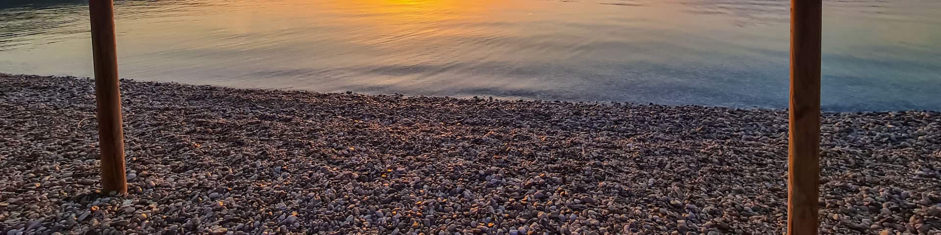 A calm lake at sunset with two thatched umbrellas on pebbled shore.