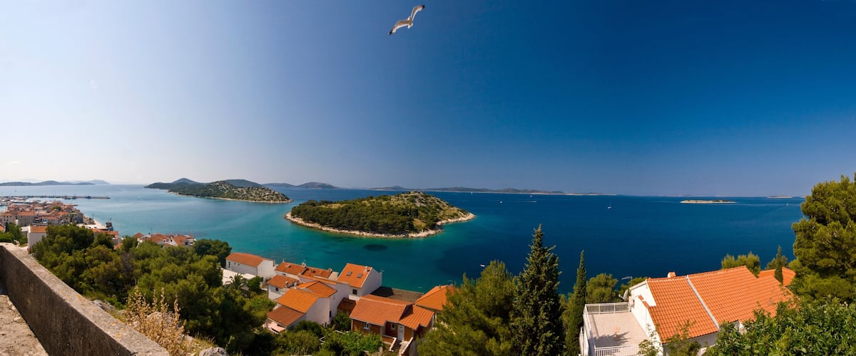 A coastal town with terracotta-roofed houses, a clear blue sea, and a small island with lush greenery.