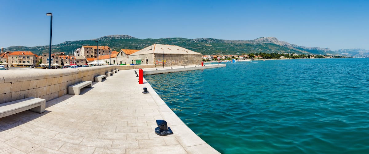 A coastal promenade with a clear view of the sea, a stone wall, and buildings with red roofs.