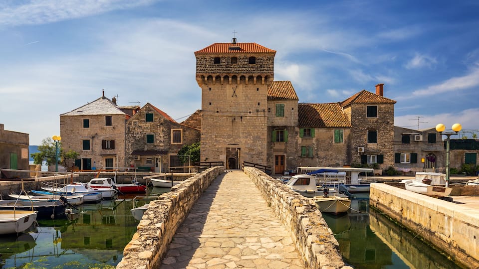 A stone pathway leading to a historic building with a tower, surrounded by boats and water.