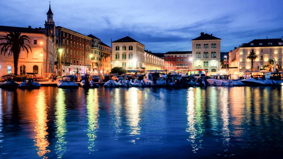 A waterfront with boats docked, buildings with illuminated facades, and a clear reflection on the water.
