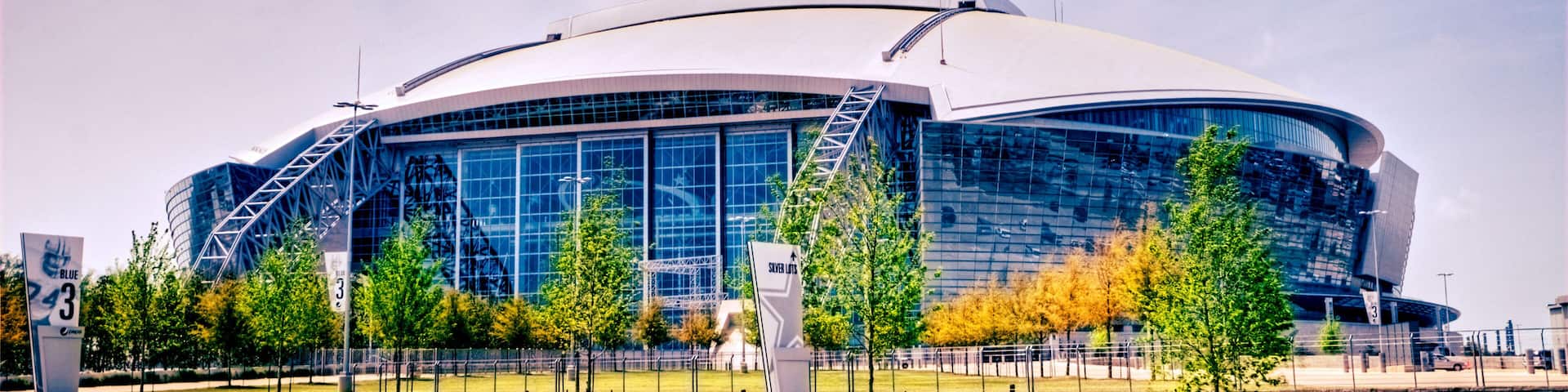 A large, modern stadium with a curved roof and glass facade, surrounded by trees and a paved walkway.