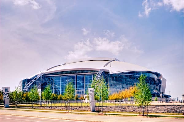 A large, modern stadium with a curved roof and glass facade, surrounded by trees and a paved walkway.