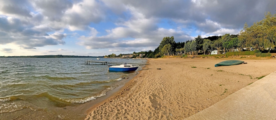 Une plage de sable avec un bateau bleu amarré sur le rivage, un chemin en béton et une rangée de maisons en arrière-plan.