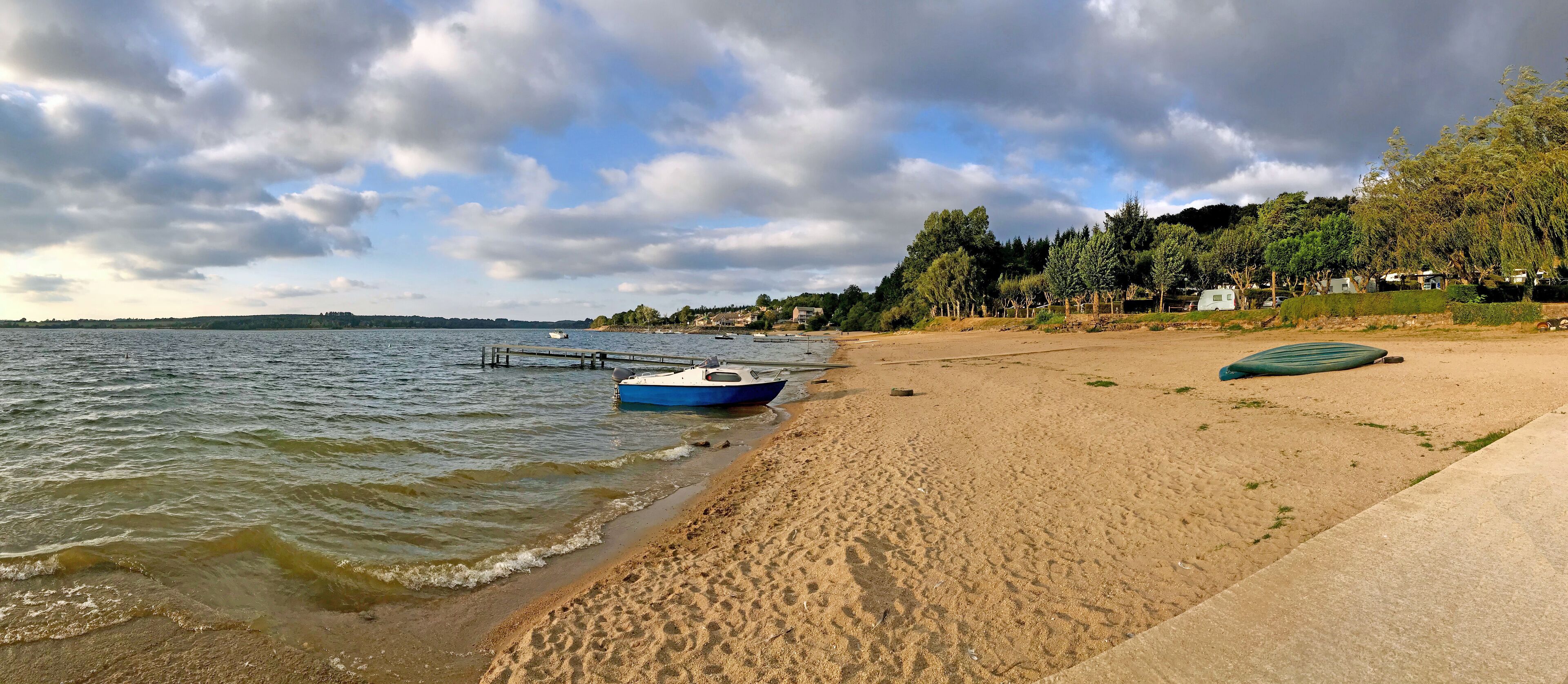 A sandy beach with a blue boat docked on the shore, a concrete path, and a row of houses in the background.