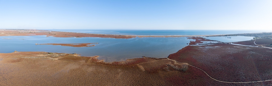 Vista aérea de uma paisagem costeira com uma grande extensão de água, áreas arenosas e uma linha de costa distante.