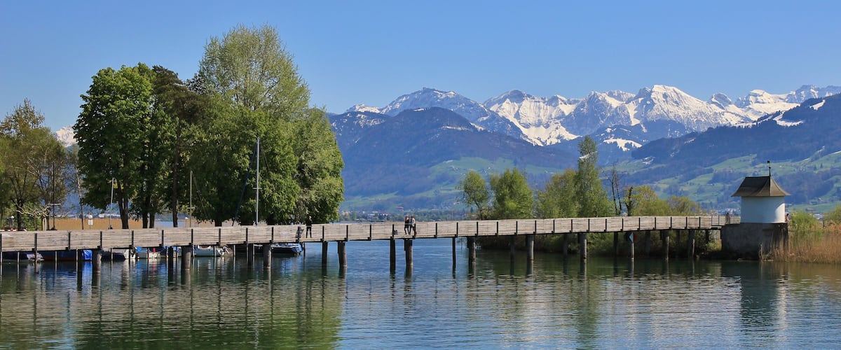 A wooden pier extending over a calm lake, with snow-capped mountains in the background.