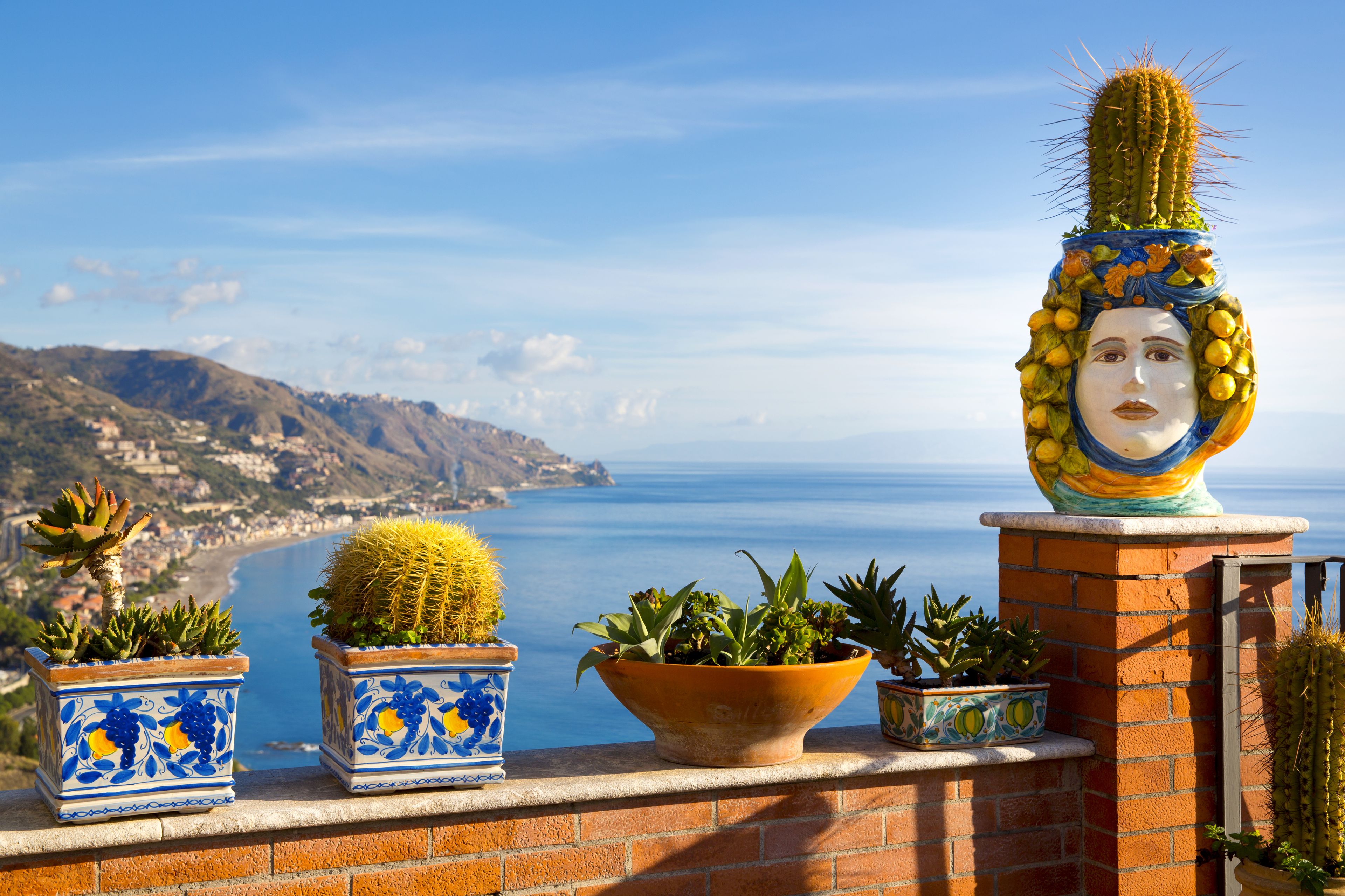 Vista costera con una escultura de cactus sobre un muro de ladrillo, plantas en macetas y un mar azul transparente.