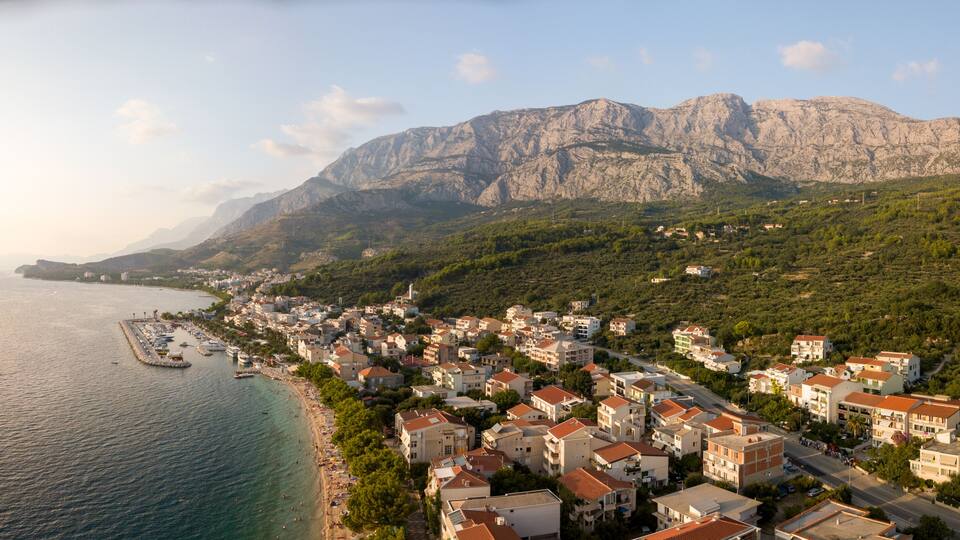 A coastal town with a marina, buildings, and a mountain range in the background.