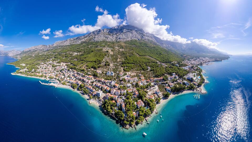 A coastal town with a mountainous backdrop, clear blue waters, and a small island with buildings.