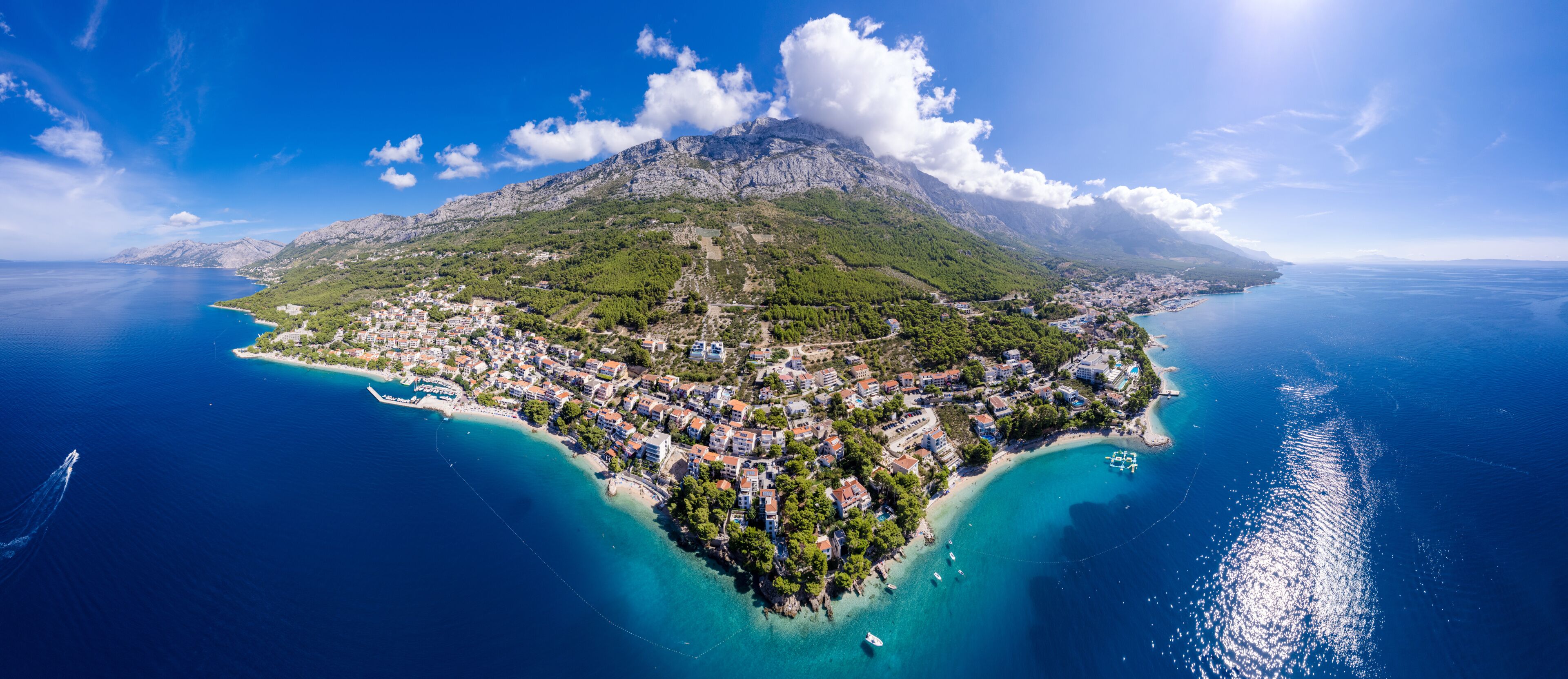 A coastal town with a mountainous backdrop, clear blue waters, and a small island with buildings.