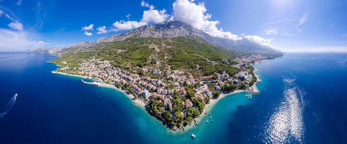 A coastal town with a mountainous backdrop, clear blue waters, and a small island with buildings.