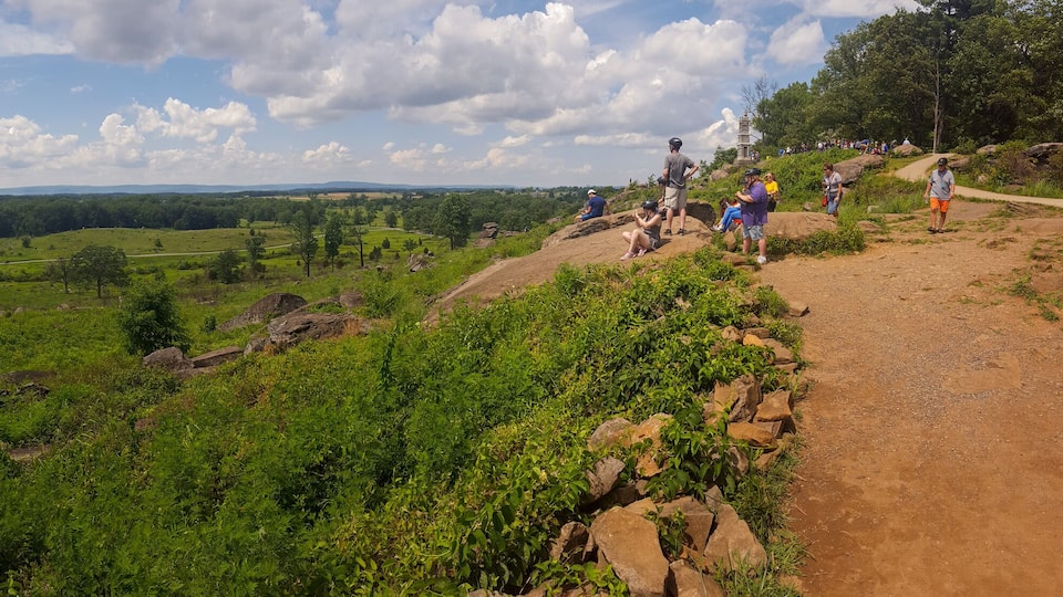 A lush green valley with a winding path, rocky outcrops, and a distant view of hills under a partly cloudy sky.