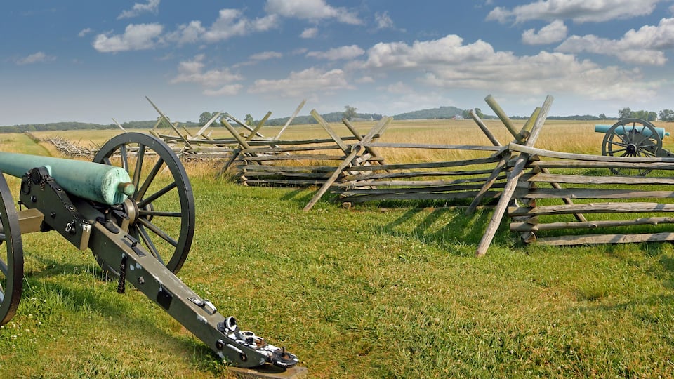 A cannon in a grassy field with wooden fences and another cannon in the background.