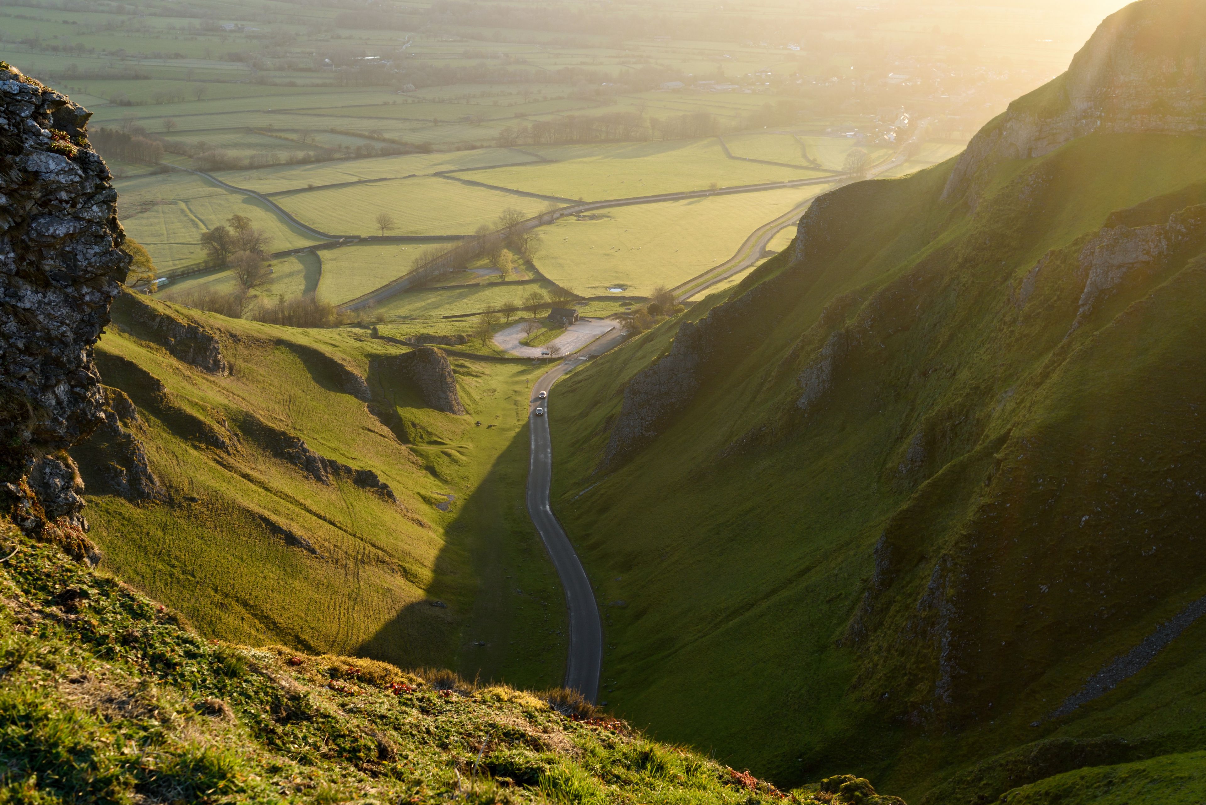 A winding road through a valley with green hills and a distant view of fields.