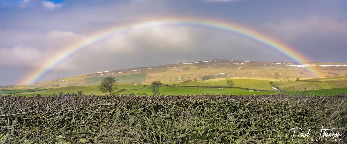 A rainbow over a hilly landscape with snow patches and a field of crops.