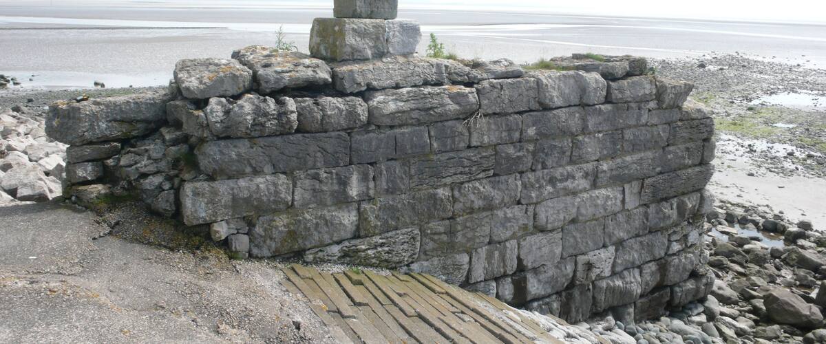 A stone wall with a standing stone on top, located near a beach with a view of the sea.