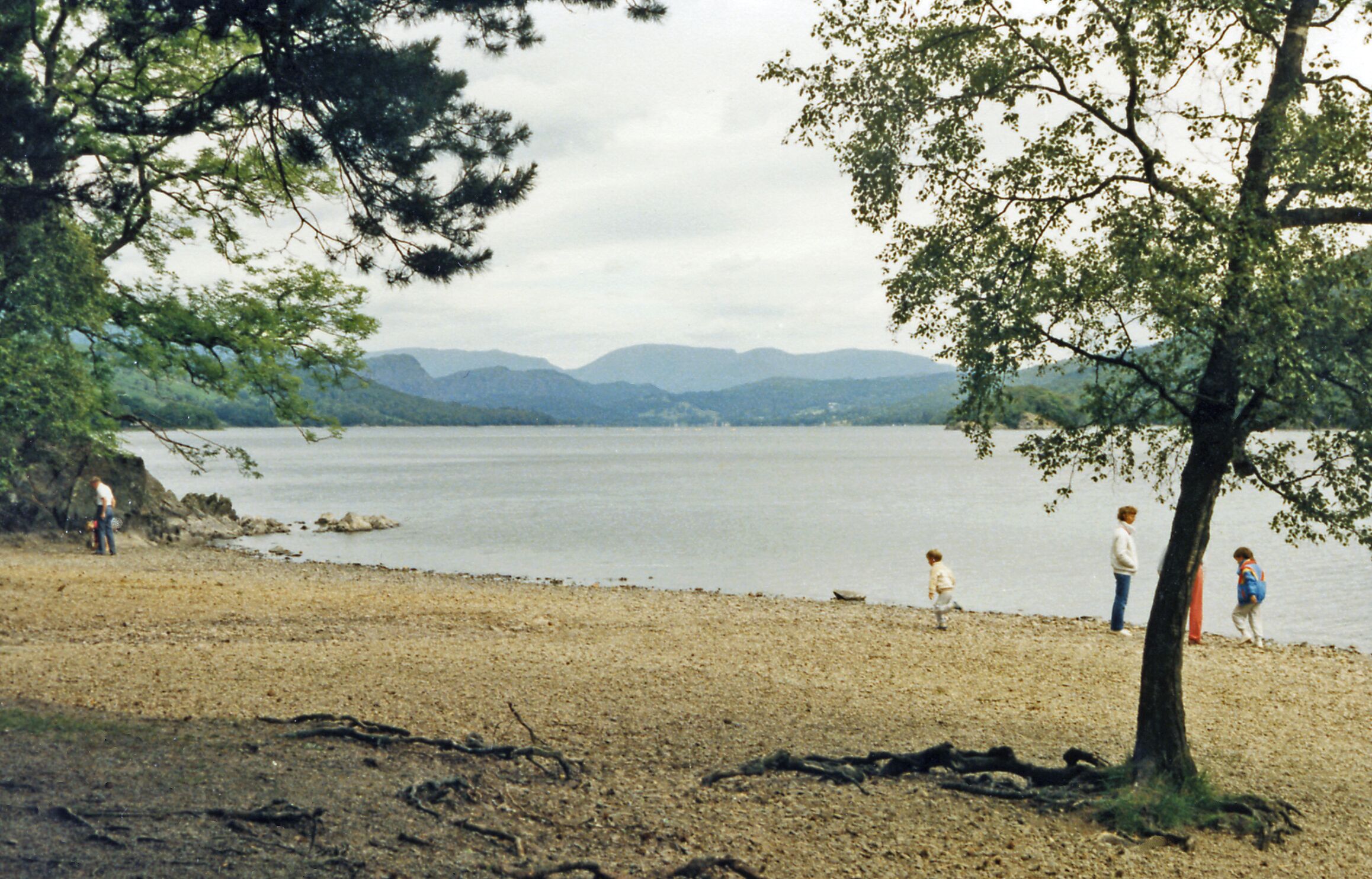 Un lago con montañas al fondo, una playa de arena y algunas personas caminando por la orilla.