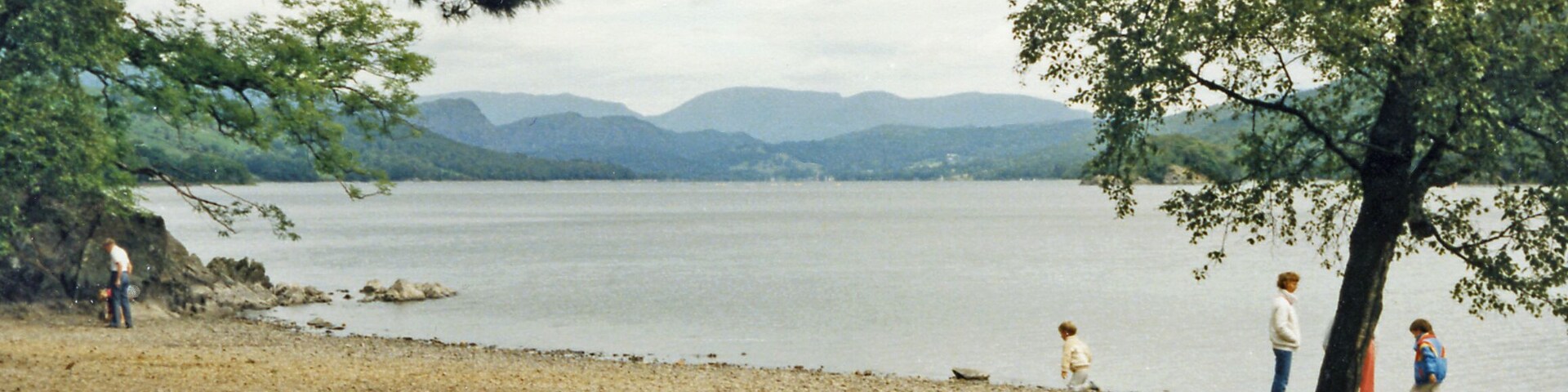 Un lago con montañas al fondo, una playa de arena y algunas personas caminando por la orilla.