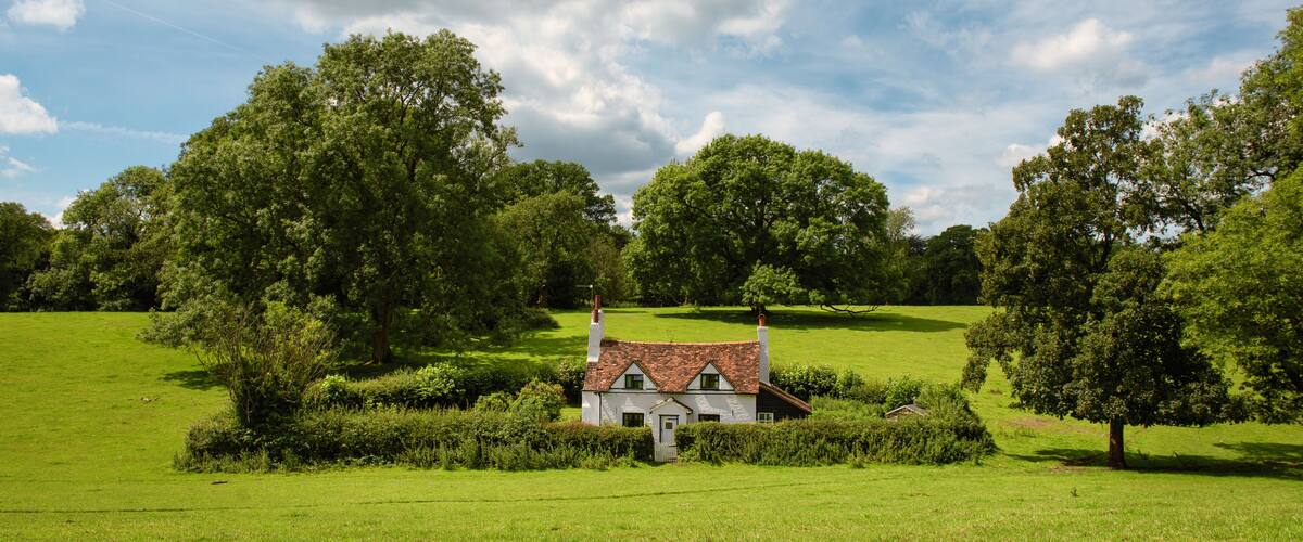 A quaint white cottage with a red-tiled roof surrounded by lush green fields and large trees.