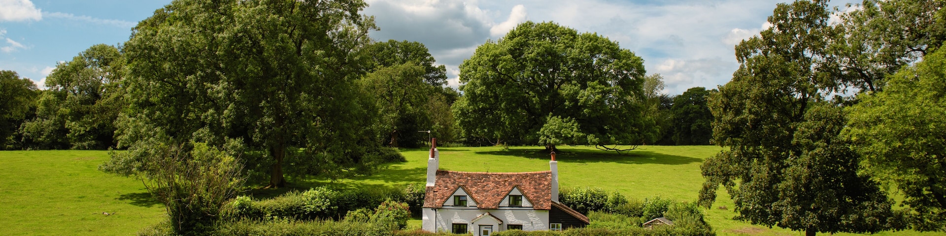 A quaint white cottage with a red-tiled roof surrounded by lush green fields and large trees.