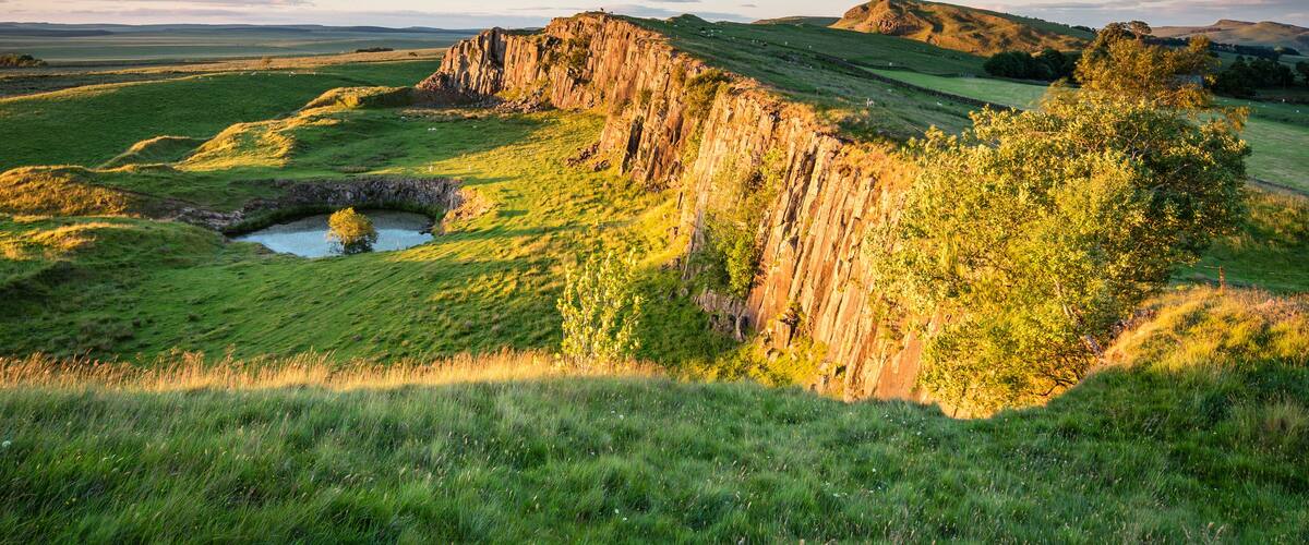 A grassy valley with a rocky cliff and a small pond.