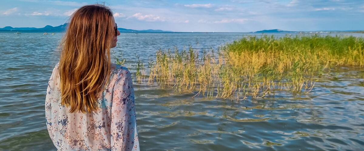Tourist woman standing at idyllic lakeshore with panoramic view of calm Balaton lake seen from tourist town Balatonmariafurdo, Hungary, Europe. Tranquil serene atmosphere. Travel destination in summer