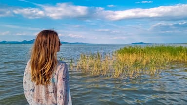 Tourist woman standing at idyllic lakeshore with panoramic view of calm Balaton lake seen from tourist town Balatonmariafurdo, Hungary, Europe. Tranquil serene atmosphere. Travel destination in summer