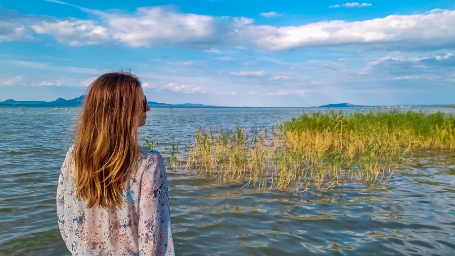 Tourist woman standing at idyllic lakeshore with panoramic view of calm Balaton lake seen from tourist town Balatonmariafurdo, Hungary, Europe. Tranquil serene atmosphere. Travel destination in summer