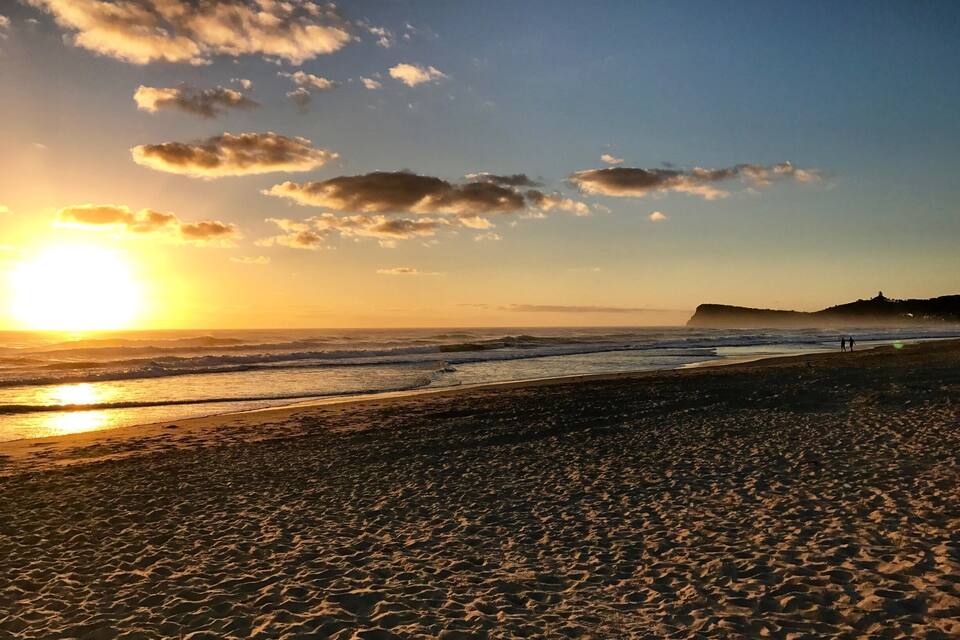 A beach at sunset with waves, a clear sky, and a distant hill.