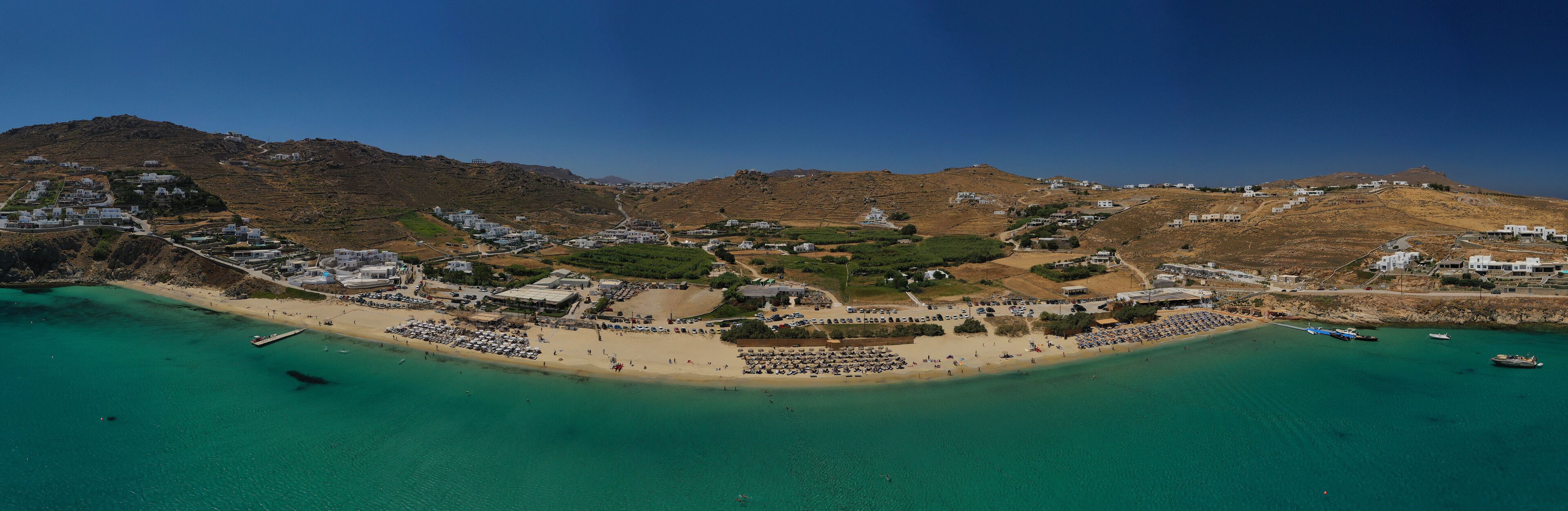 A coastal town with a beach, clear turquoise waters, and hilly terrain in the background.