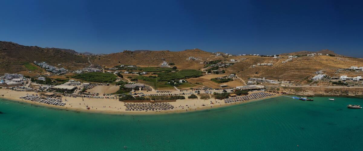 A coastal town with a beach, clear turquoise waters, and hilly terrain in the background.