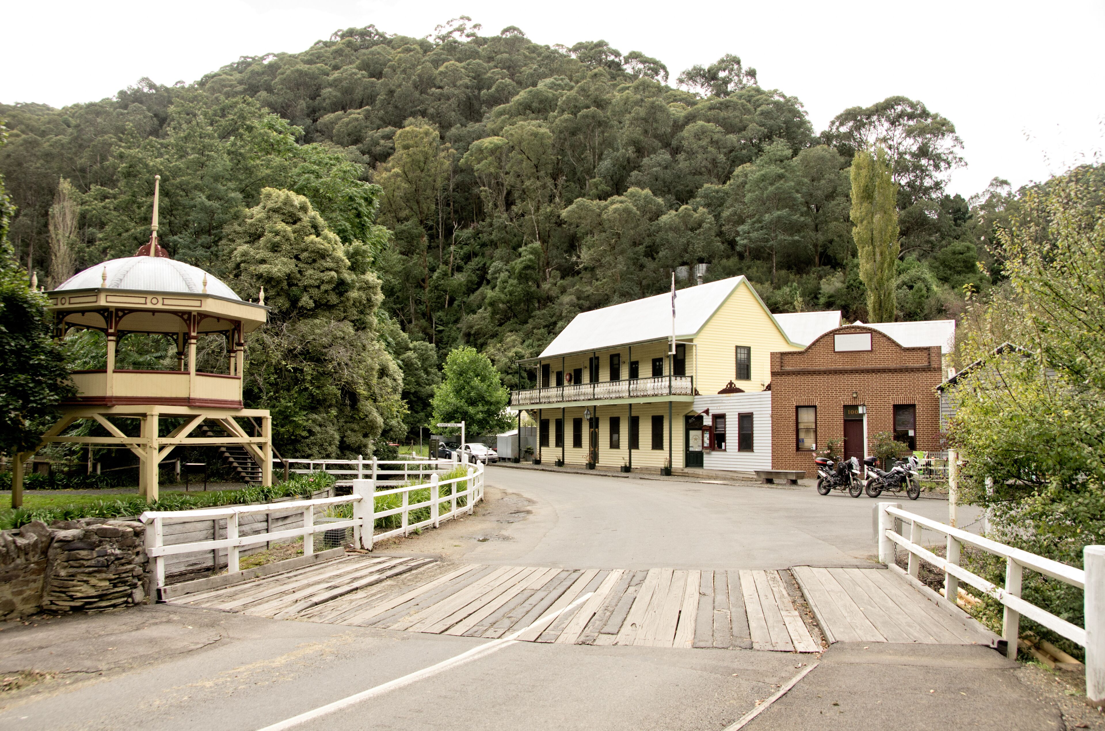 A wooden bridge, a gazebo, and buildings with a hill in the background.