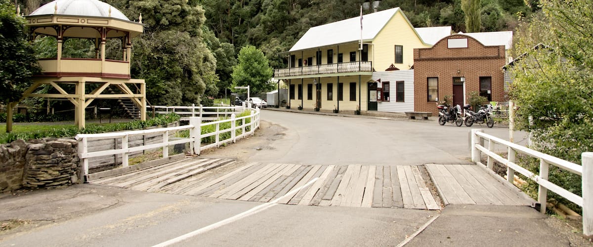 A wooden bridge, a gazebo, and buildings with a hill in the background.