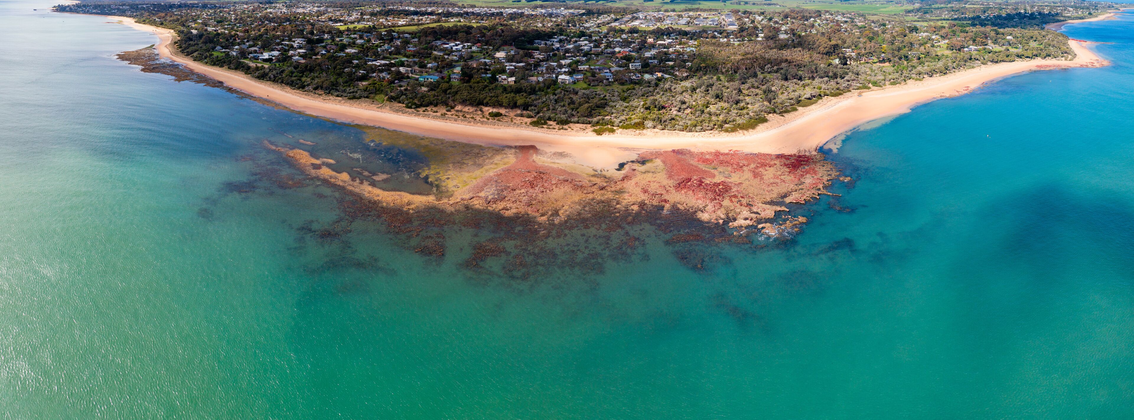 An aerial view of a coastal area with a beach, a forested peninsula, and clear blue waters.