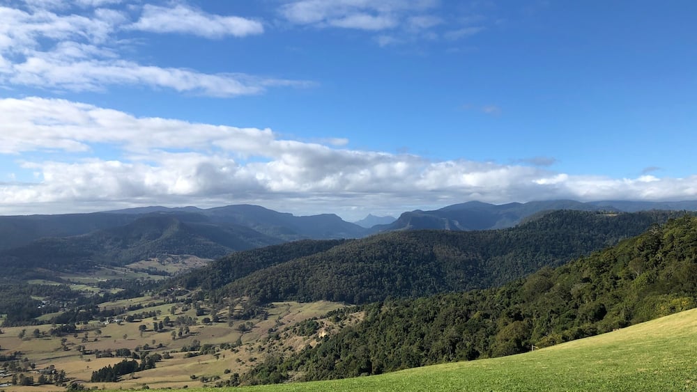 A mountainous landscape with rolling hills, dense forests, and a clear blue sky with scattered clouds.
