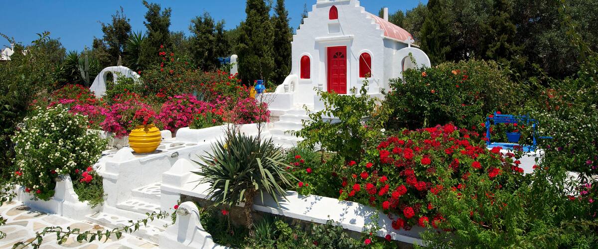 A white church with red doors and a bell tower surrounded by lush greenery and colorful flowers.