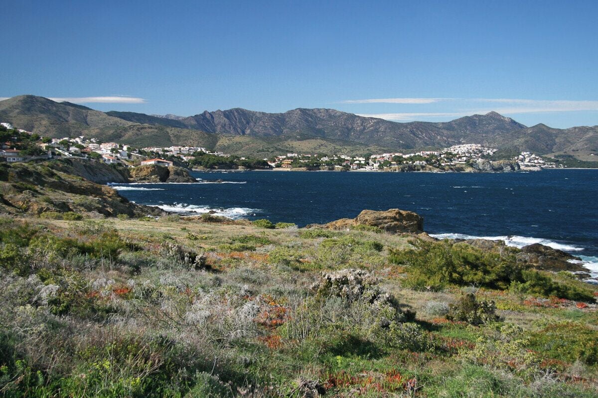 A coastal landscape with rugged mountains, a rocky shoreline, and a small settlement with white buildings.
