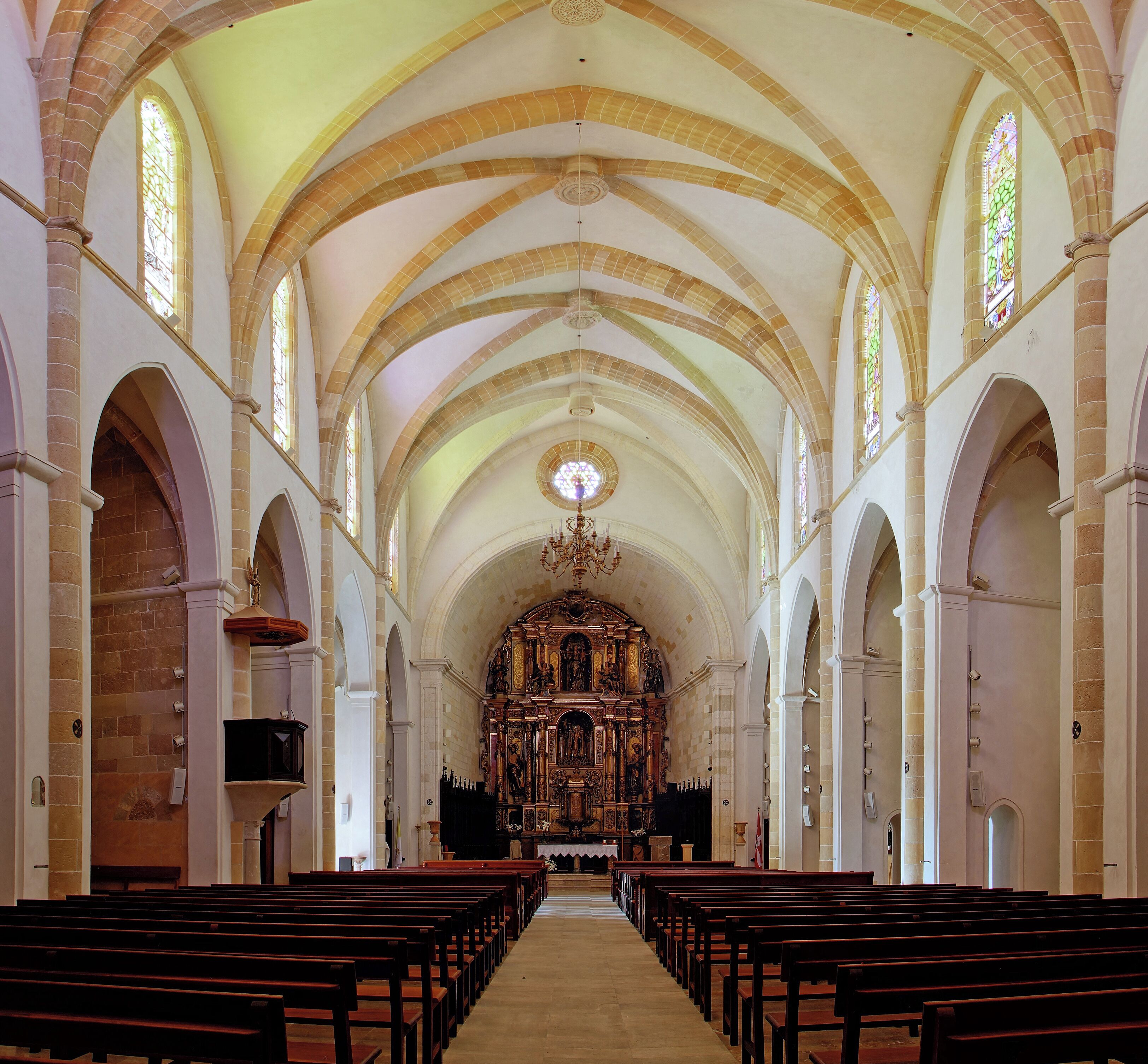 Interior de una iglesia con techos abovedados, vidrieras de colores y hileras de bancas.