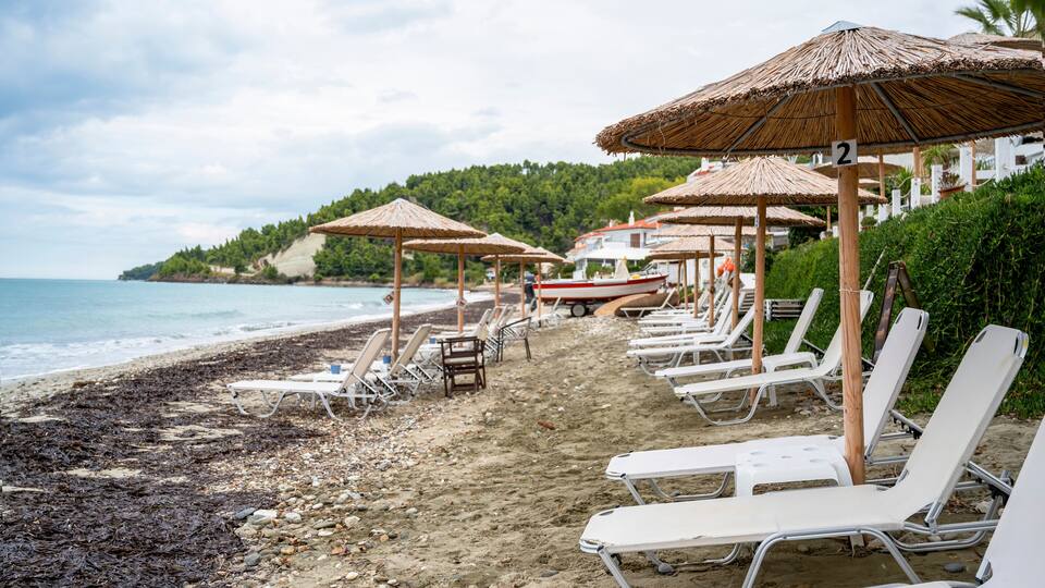 A beach with rows of sun loungers and thatched umbrellas, a boat, and a forested hillside in the background.