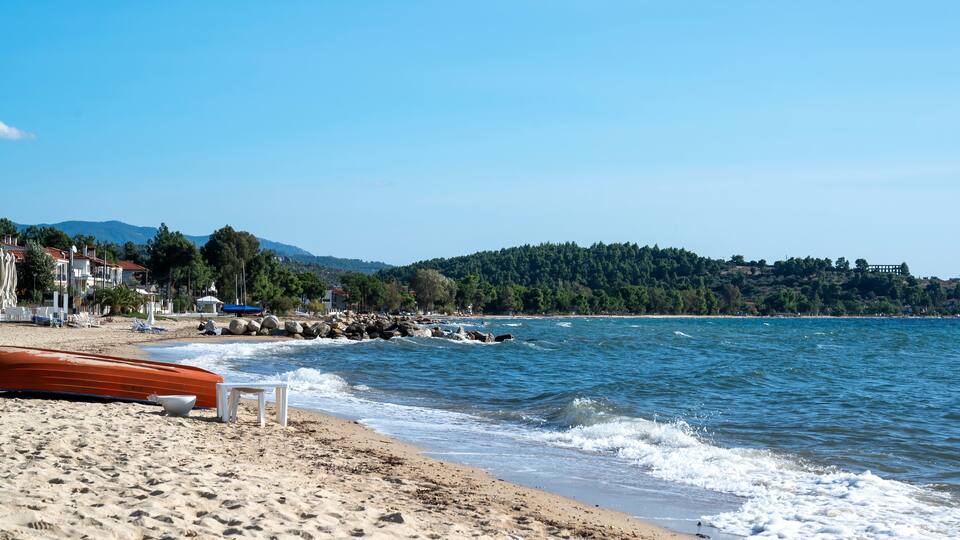 A beach with a rocky shoreline, a row of houses, and a forested hill in the background.