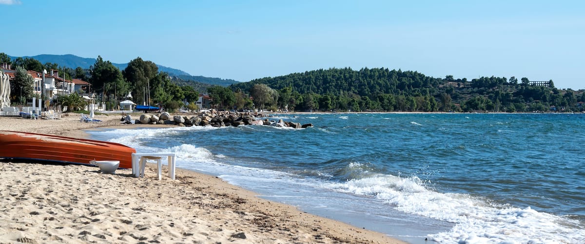 A beach with a rocky shoreline, a row of houses, and a forested hill in the background.