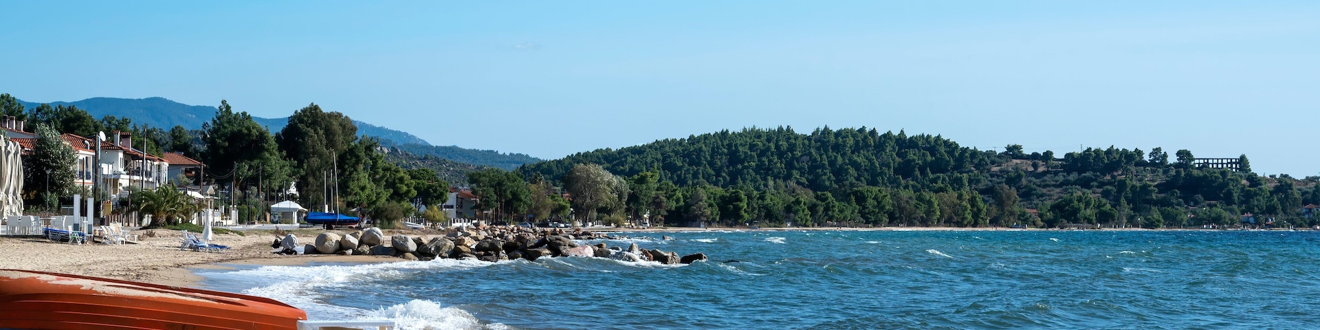 A beach with a rocky shoreline, a row of houses, and a forested hill in the background.