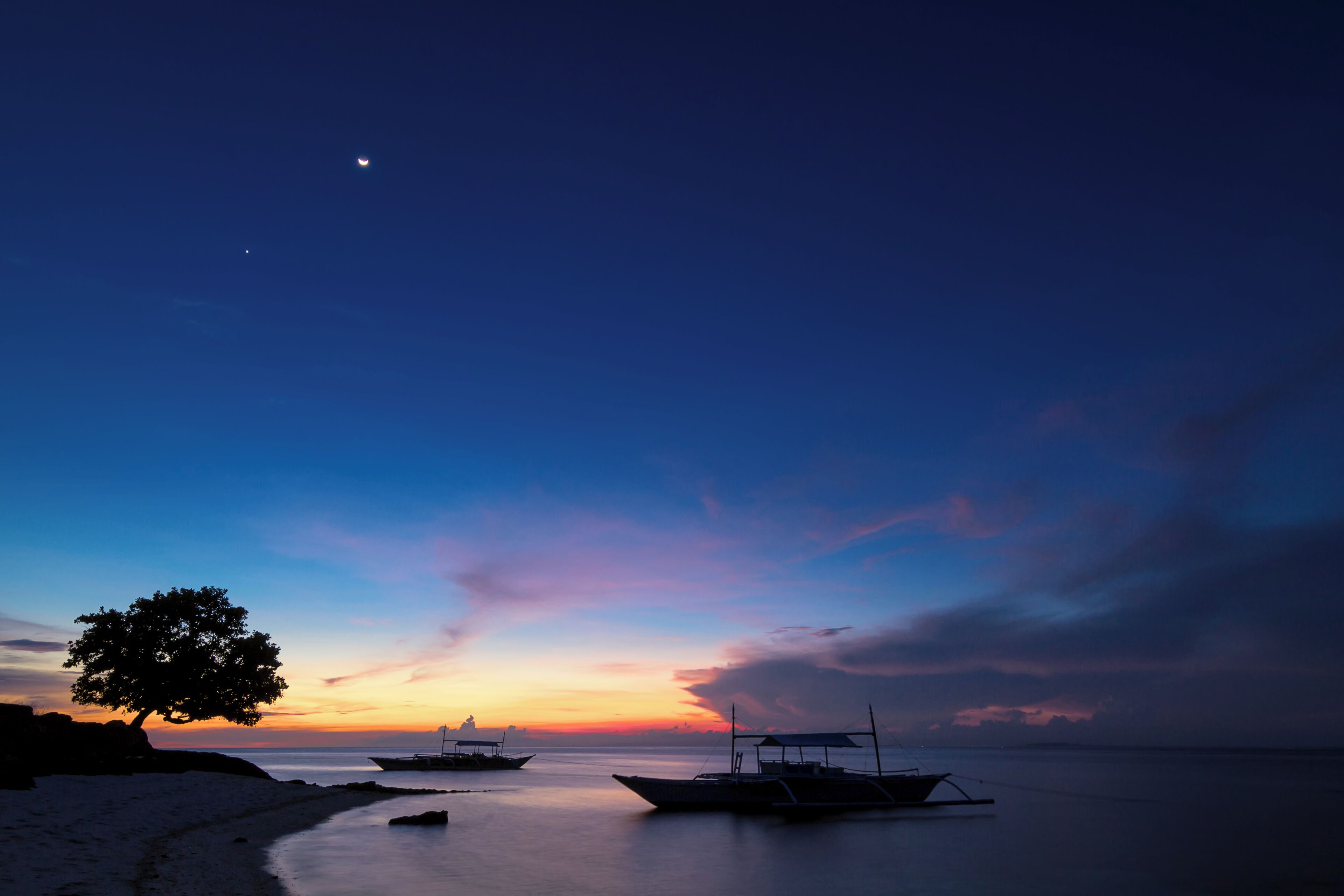 A beach at sunset with boats and a tree silhouetted against the sky.