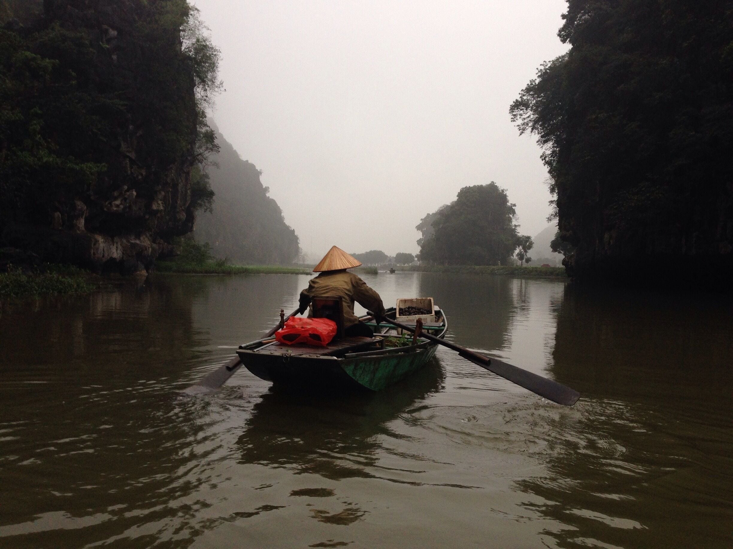 A person in a boat with a conical hat paddling through a misty river surrounded by dense forest.