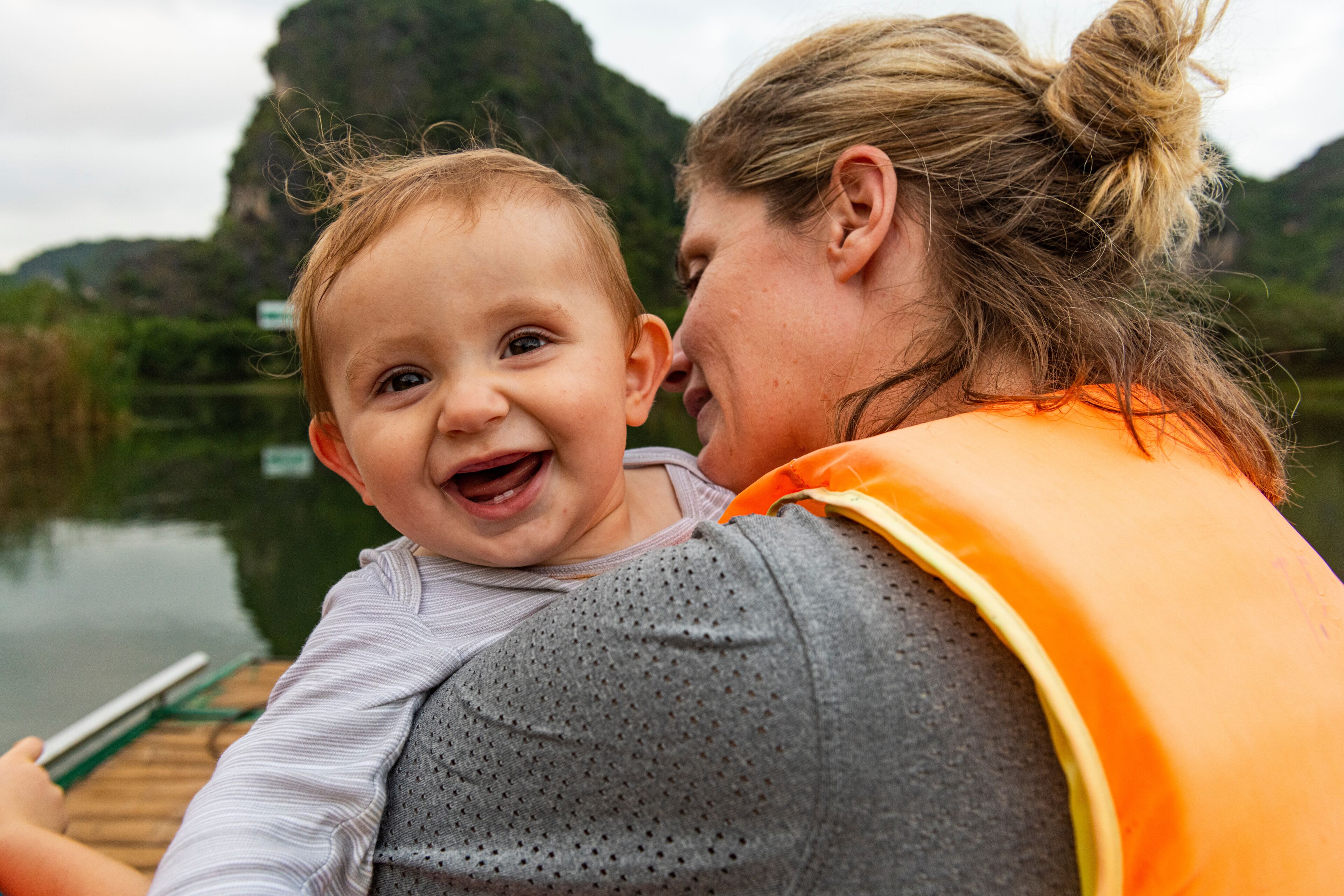 A person holding a baby by a calm river with mountains in the background.