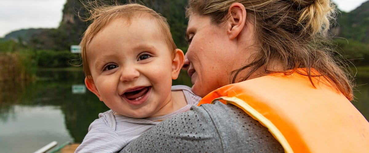 A person holding a baby by a calm river with mountains in the background.