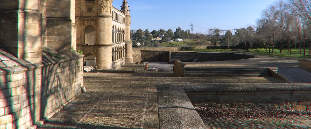 A historic stone building with a clock tower, a paved courtyard, and a park with trees in the background.