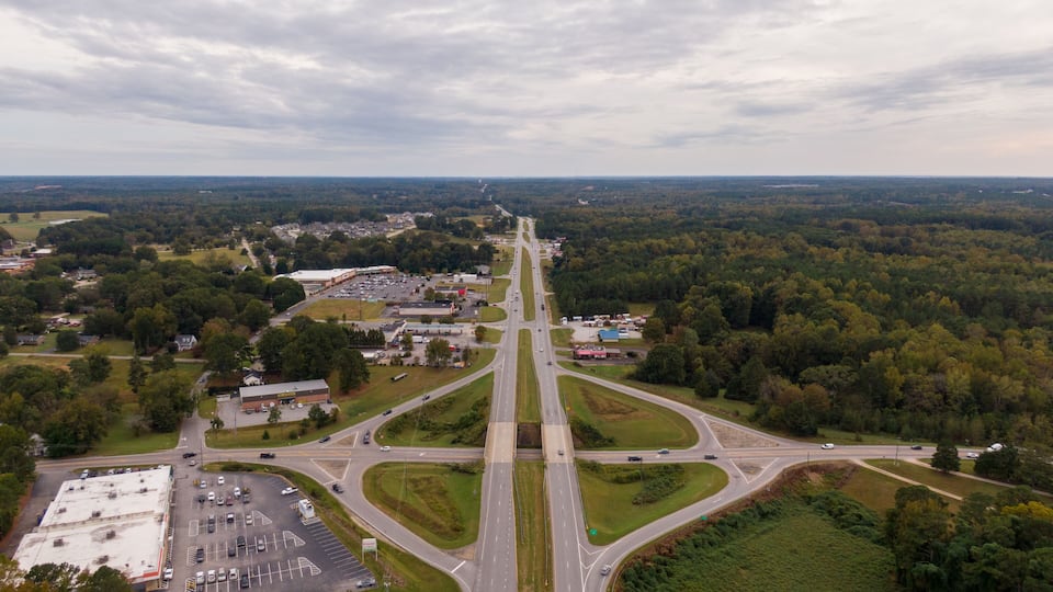 An aerial view of a highway interchange surrounded by dense forests and a mix of commercial and residential buildings.