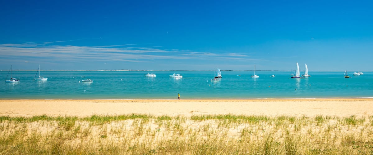 A beach with clear turquoise water, several sailboats, and a sandy shore.